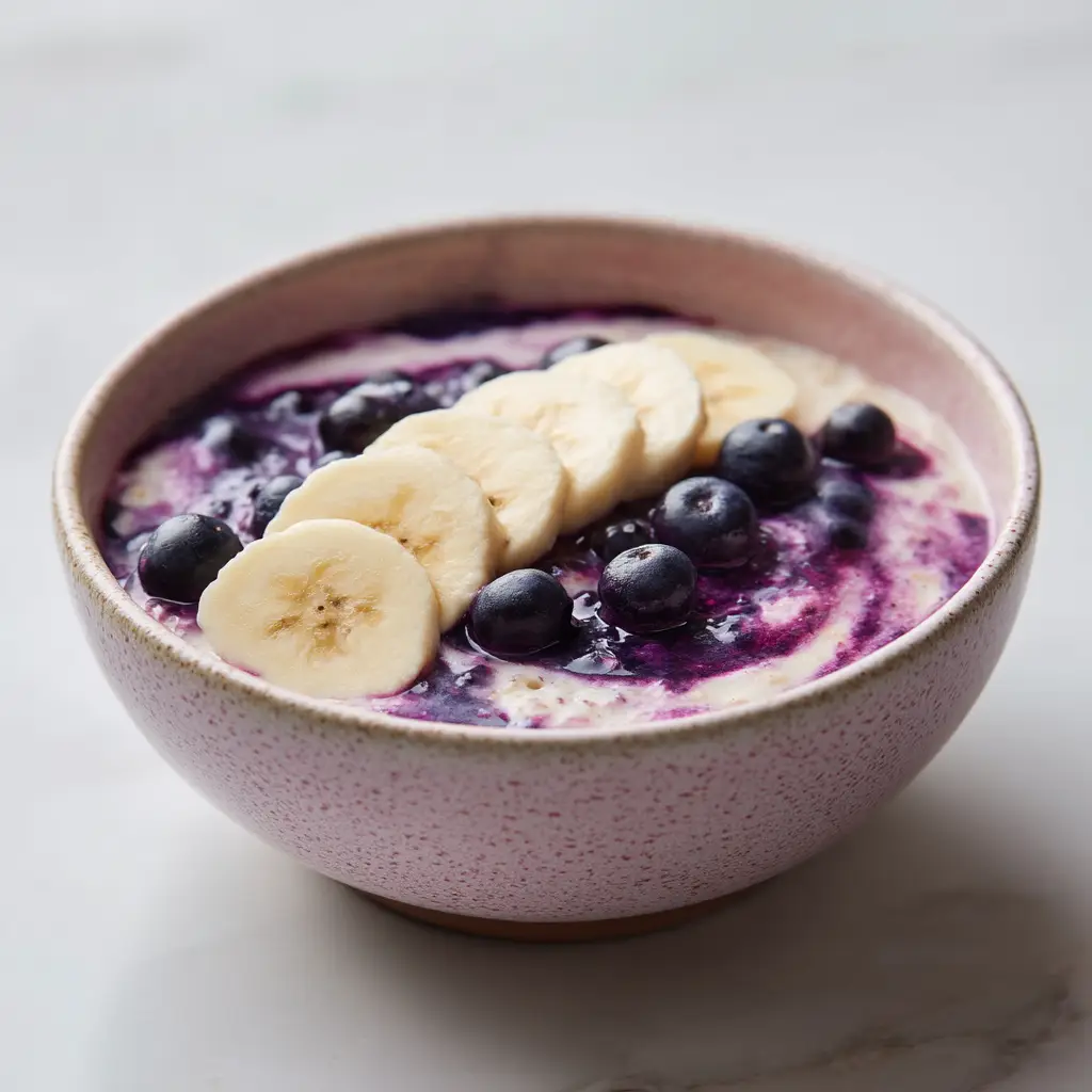 A close-up shot of healthy blueberry banana overnight oats in a pink ceramic bowl, ready to be eaten. The texture is thick and creamy.