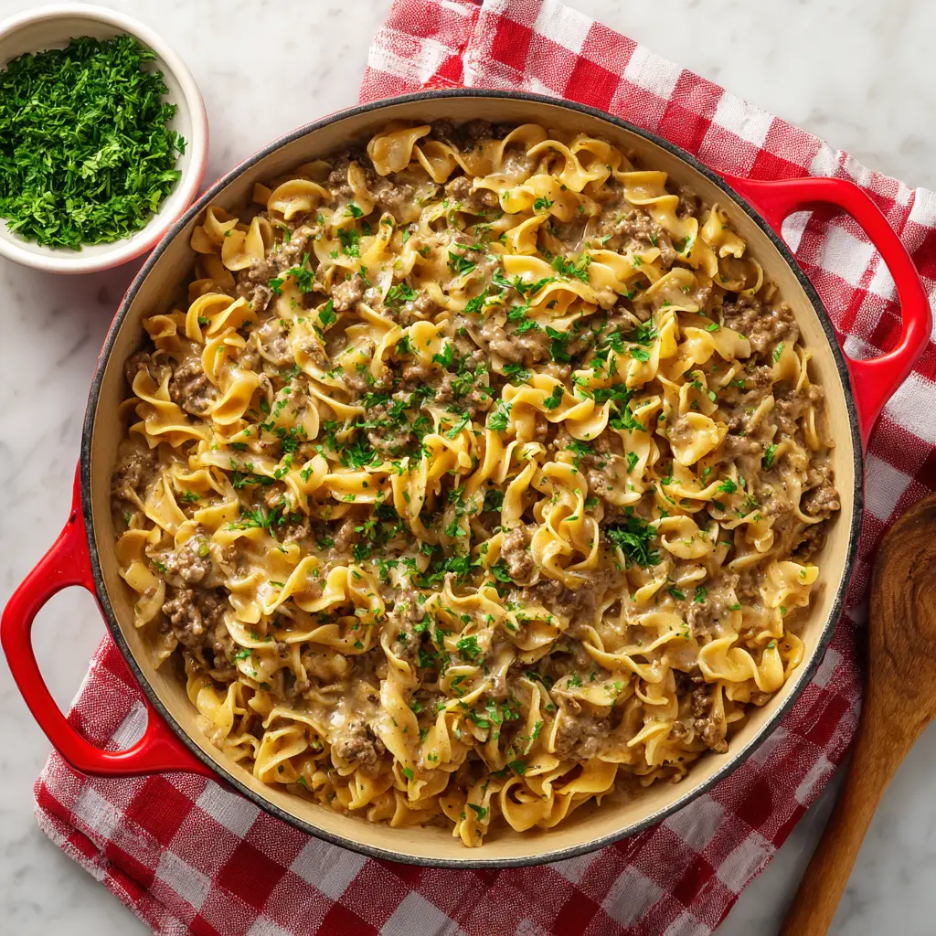 A rustic red and white checkered cloth under a red cast-iron skillet filled with creamy hamburger stroganoff, garnished with fresh parsley.