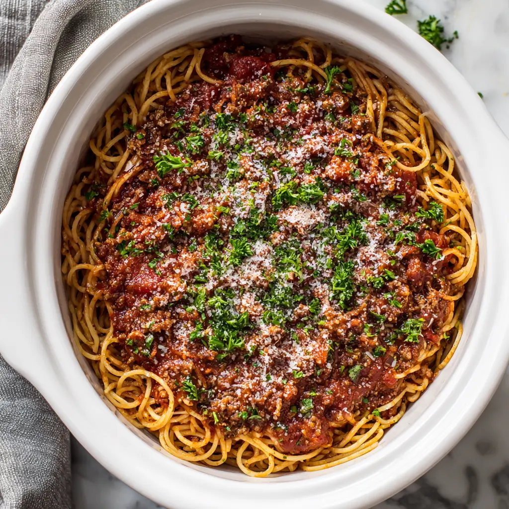 Rustic plating of Crockpot Spaghetti filling a white ceramic crockpot, topped with grated parmesan cheese, bright green chopped parsley, with a grey dish towel on the left.