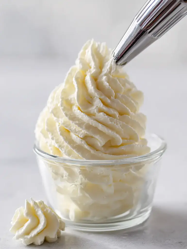A close-up photo of velvety mascarpone frosting being piped into a clear ramekin using a metal star tip. (Mascarpone Frosting Recipe)
