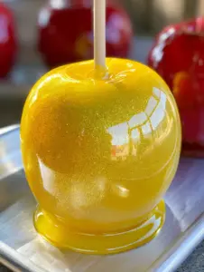 Close up photo of a yellow glossy candy apple with gold glitter and a wooden stick. (Pineapple Lemonade Candy Apples)