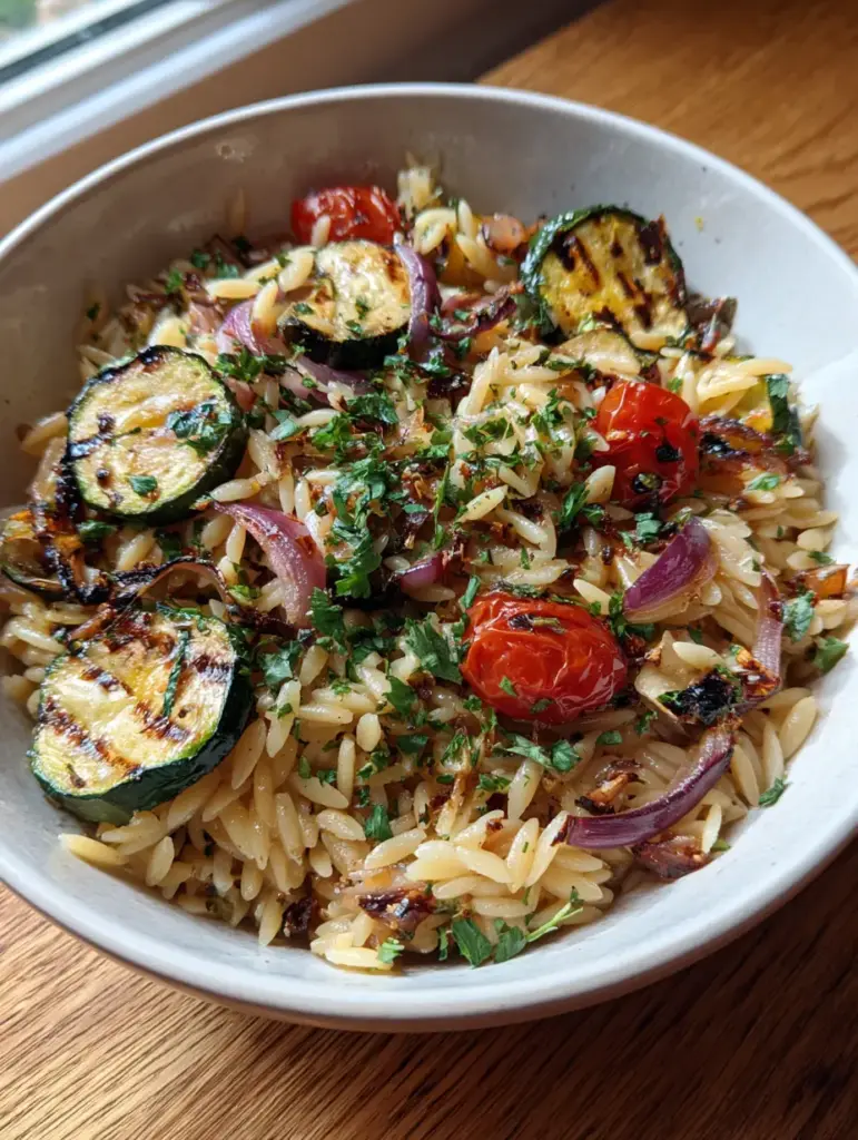 A white ceramic bowl of orzo salad with caramelized red onions and fresh parsley garnish under natural daylight. (Roasted Vegetable Orzo Salad)