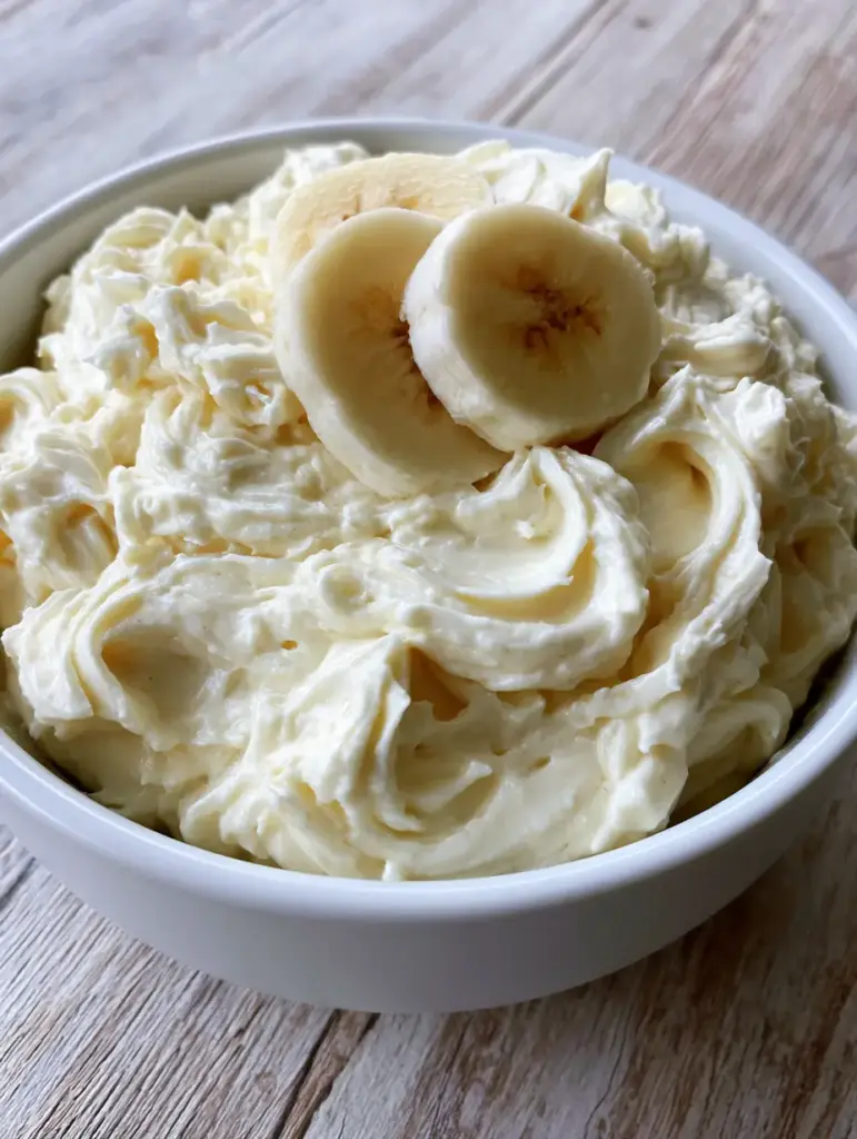 Close-up of whipped banana pudding frosting with billowy peaks in a white ceramic bowl. (Banana Pudding Frosting)