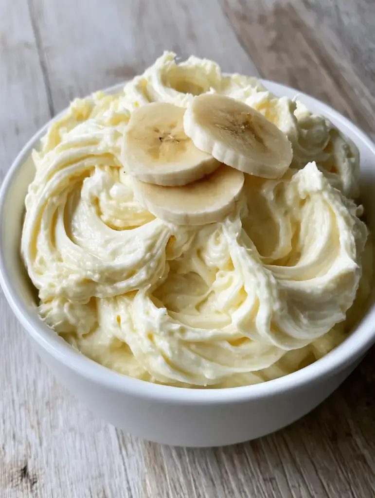 Whipped banana pudding frosting garnished with three thin slices of fresh banana on a wooden table. (Banana Pudding Frosting)