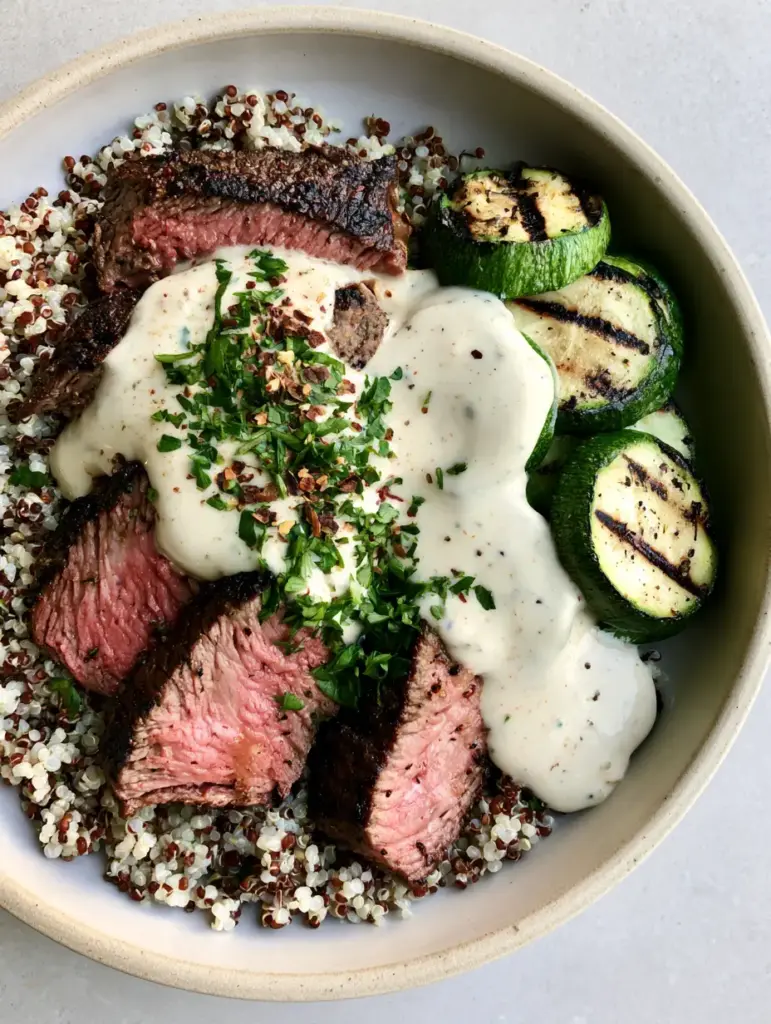 High-angle shot of the Grilled Steak Bowl highlighting the grilled zucchini rounds and the thick white garlic cream sauce.